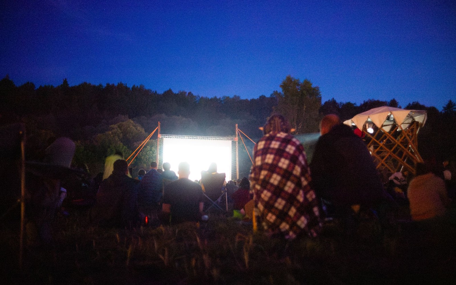 People wrapped in blankets watch a glowing outdoor movie screen at dusk