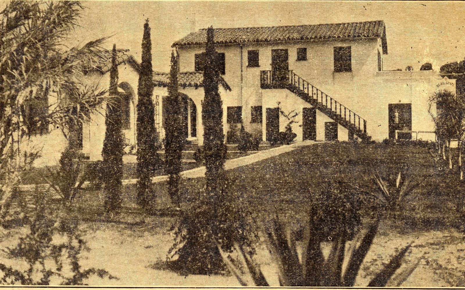 Old photograph of a Spanish-style villa with an exterior staircase, framed by tall cypress trees