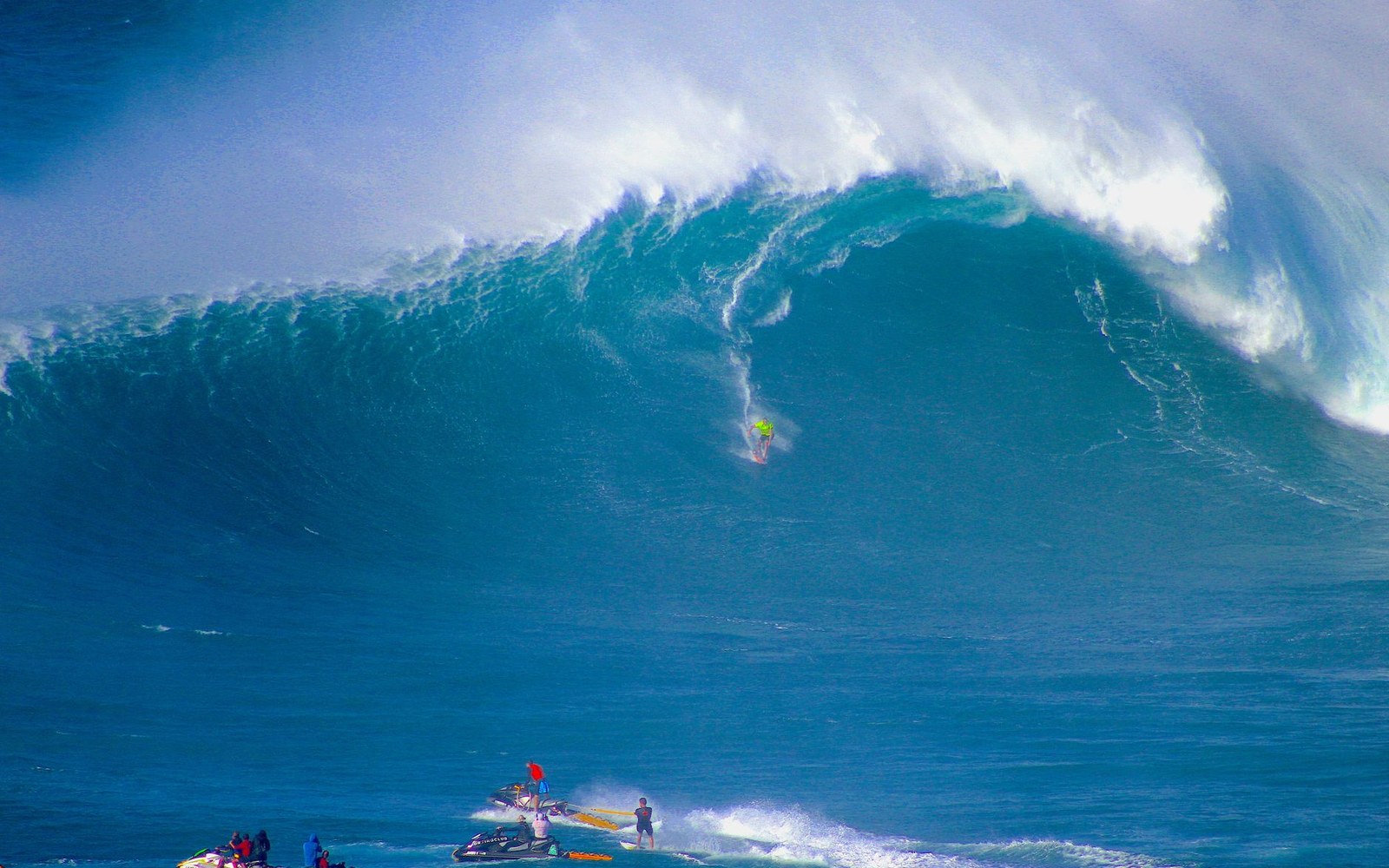 A surfer descends a massive curling wave as jet skis wait near the base