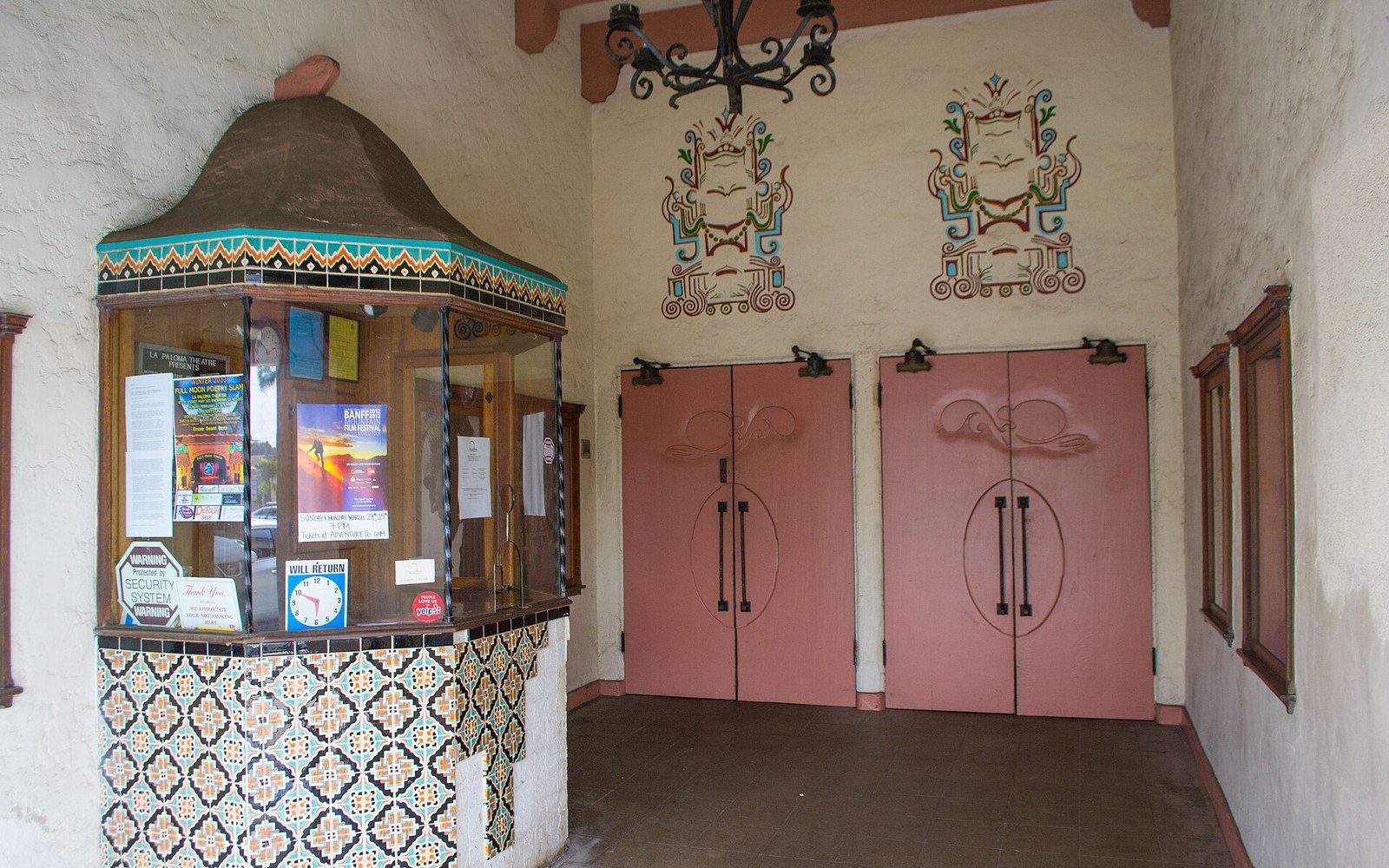 Tiled ticket booth beside pink double doors under painted murals at a Spanish-style theater entrance