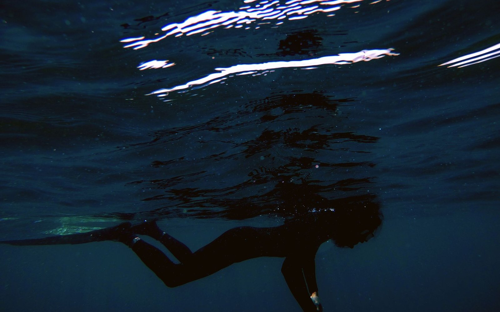 Silhouette of a swimmer gliding underwater beneath rippling, light-streaked ocean surface
