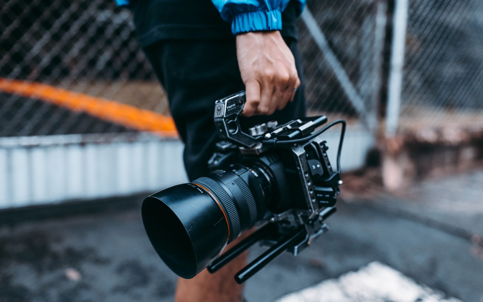 A person carries a rigged cinema camera with lens hood by its handle outdoors