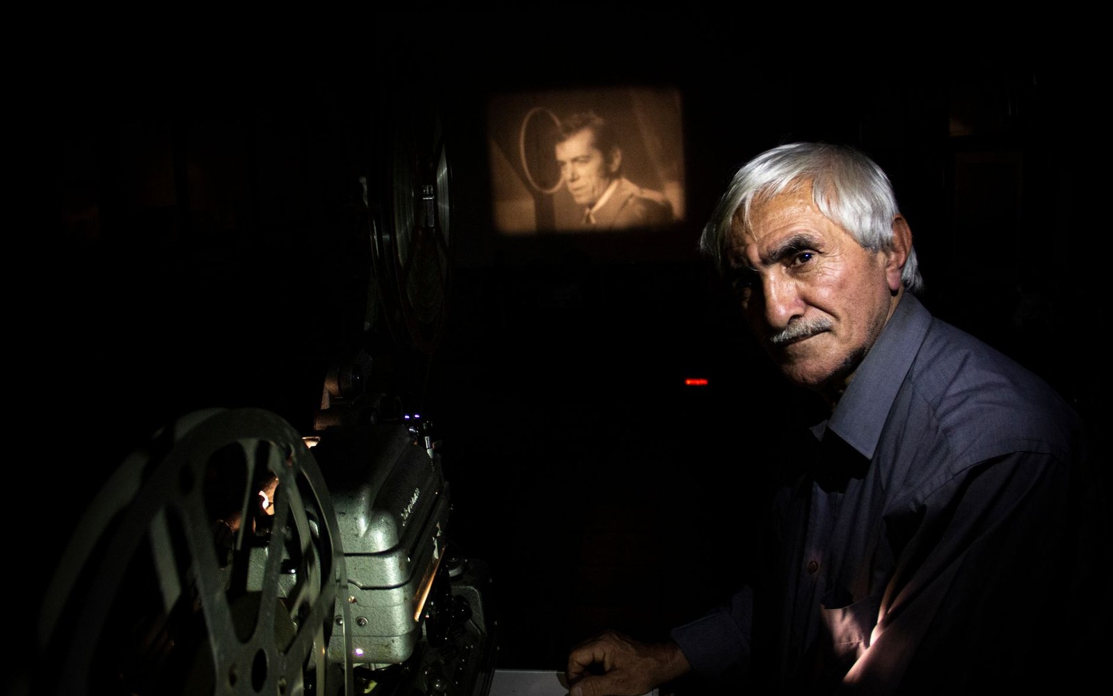 An adult projection booth operator at work, projector running, light beam visible through the port
